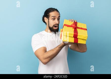 Portrait of handsome man with beard wearing white T-shirt unpacking present, looking inside box with shocked and scared expression, birthday gift. Indoor studio shot isolated on blue background. Stock Photo