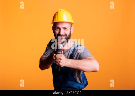 Cheerful construction worker holding power drill gun and posing in studio, looking at camera. Craftsman builder using drilling nail tool wearing uniform and hardhat, renovating service. Stock Photo