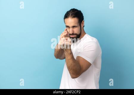 Portrait of assertive dangerous man with beard wearing white T-shirt pointing fingers at target imitating weapon making shot, terrorism. Indoor studio shot isolated on blue background. Stock Photo