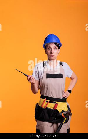 Inexperienced female builder doing renovation project and holding screwdriver, wearing work uniform in studio. Industrial construction worker using screw tool for nails, professional handywoman. Stock Photo