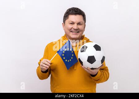 Satisfied middle aged man holds in hands flag of europe union and football ball, supporting favourite team on championship, wearing urban style hoodie. Indoor studio shot isolated on white background. Stock Photo