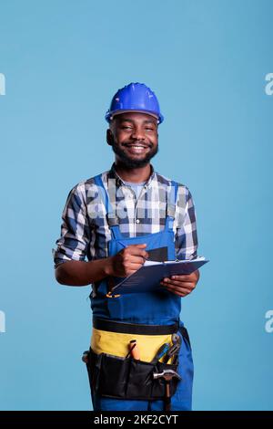 Portrait of construction contractor in hard hat and coveralls standing in front of a blue background holding a clipboard reading a report. Optimistic builder reviewing construction paperwork in studio shot. Stock Photo