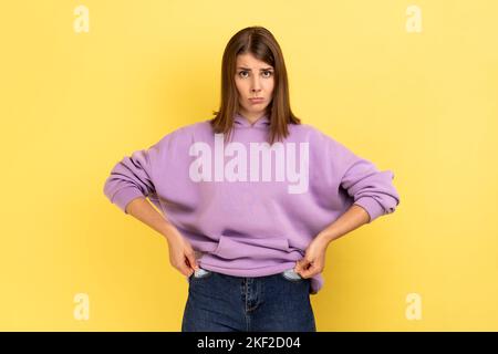 Unhappy poor woman turning empty pockets inside out and looking frustrated by overspend, lack of money, financial crisis, wearing purple hoodie. Indoor studio shot isolated on yellow background. Stock Photo