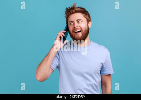 Portrait of excited handsome bearded man standing, talking smartphone, making call and having pleasant conversation with friend or wife. Indoor studio shot isolated on blue background. Stock Photo