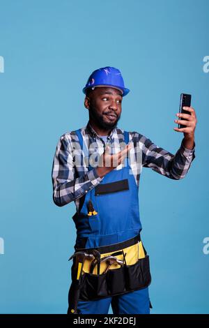 African american contractor talking on video call with business partner wearing coveralls against blue background in studio shot. Construction worker talking pleasantly on cell phone, wearing hard hat and tool belt. Stock Photo