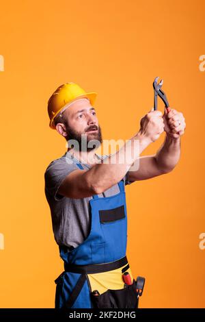 Young foreman builder looking focused at pair of pliers, using construction tools over yellow background. Male renovation expert or carpenter working with pliers for refurbishment project. Stock Photo