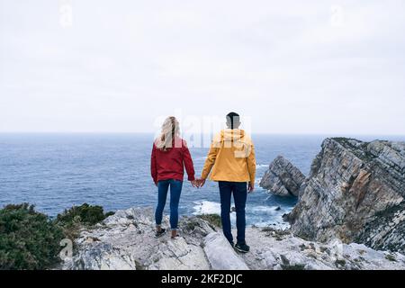 latin boy and caucasian girl in red and yellow jacket blue pants and sneakers standing holding hands looking at the horizon, cabo de penas, spain Stock Photo