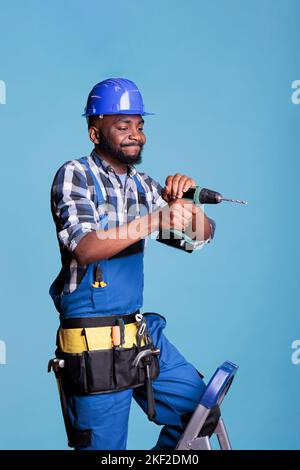 African american constructor using electric drill for screwing nails, working with engineering machine in construction renovation. Builder drilling with nail gun for repair, on blue background. Stock Photo