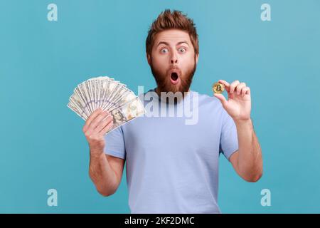 Portrait of amazed surprised handsome bearded man showing dollar banknotes and bitcoin, digital money, electronic commerce, expressing astonishment. Indoor studio shot isolated on blue background. Stock Photo