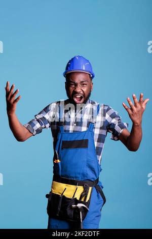 Outraged employee handyman in protective helmet outstretched hands scream isolated on blue background. Instruments accessories for construction and renovation, studio shot. Stock Photo