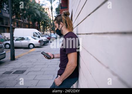 young caucasian man with long hair collected standing with his left hand in his pants pocket wearing a black t-shirt leaning against a wall looking at Stock Photo