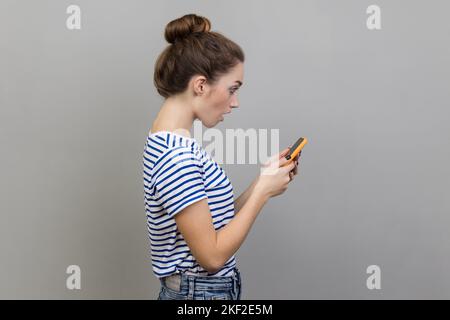 Side view of beautiful astonished woman blogger wearing striped T-shirt using smart phone, looking at device screen, sees shocking content. Indoor studio shot isolated on gray background. Stock Photo