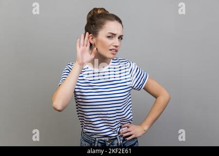 Portrait of nosy woman wearing striped T-shirt listening attentively holding hand near ear to hear confidential talk, hard to understand. Indoor studio shot isolated on gray background. Stock Photo