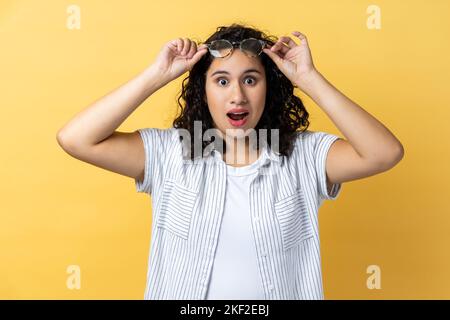 Portrait of surprised astonished woman with dark wavy hair standing raised eyeglasses and looking at camera with big eyes, sees something shocked. Indoor studio shot isolated on yellow background. Stock Photo