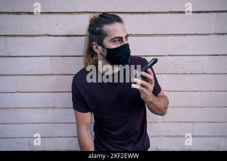 caucasian young man with long hair standing leaning against the wall talking on the smartphone wearing face mask Stock Photo