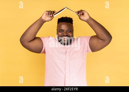 Portrait of smiling man wearing pink shirt holding opened book on head, looking at camera with smile, expressing positive emotions. Indoor studio shot isolated on yellow background. Stock Photo