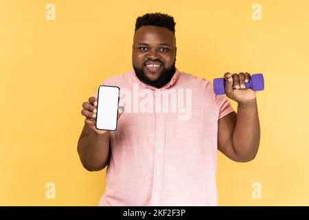 Portrait of happy joyful bearded man wearing pink shirt standing holding dumbbell and smart phone with empty display for advertisement. Indoor studio shot isolated on yellow background. Stock Photo