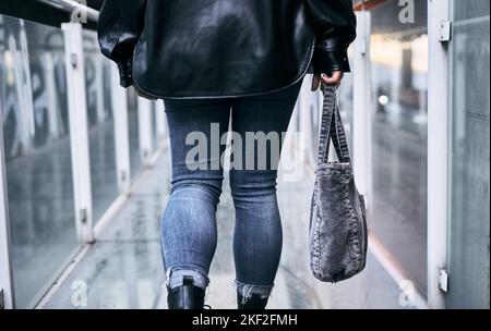 closeup legs in blue jeans of latina girl from the back walking down the glass walkway carrying a bag on the right hand side Stock Photo