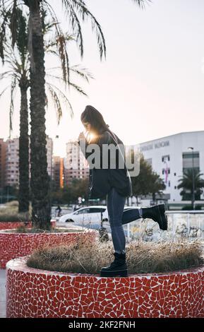 young latin brunette woman with long hair glasses blue pants standing on one leg in small garden next to palm trees in urban park Stock Photo