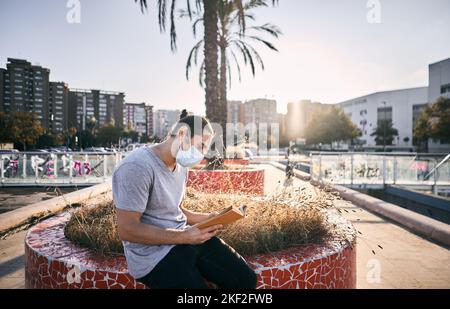 caucasian guy with long hair collected gray t-shirt face mask sitting in the urban park reading a book Stock Photo