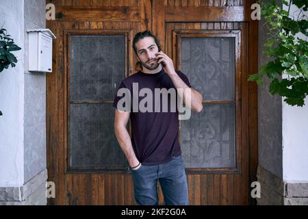 bearded caucasian young man talking on his smartphone near the wooden door and mailbox Stock Photo