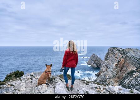 caucasian girl with a big dog sitting on a leash contemplating the beauty of the sea waves and the horizon from the big rocks on a cloudy day, cabo de Stock Photo