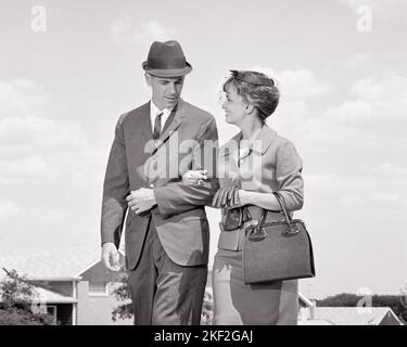 1960s MAN AND WOMAN WALKING DOWN SUBURBAN STREET TALKING ARM IN ARM MAN IN BUSINESS SUIT WOMAN IN SUIT HAT WITH GLOVES & HANDBAG - s14214 HAR001 HARS FEMALES MARRIED SPOUSE HUSBANDS HEALTHINESS COPY SPACE FRIENDSHIP HALF-LENGTH LADIES PERSONS MALES B&W PARTNER SUIT AND TIE HAPPINESS AND ARM IN ARM EXTERIOR PRIDE TO CONNECTION STYLISH SUNDAY BEST POCKETBOOK COOPERATION MID-ADULT MID-ADULT MAN MID-ADULT WOMAN TOGETHERNESS WIVES & BLACK AND WHITE CAUCASIAN ETHNICITY HAR001 OLD FASHIONED Stock Photo
