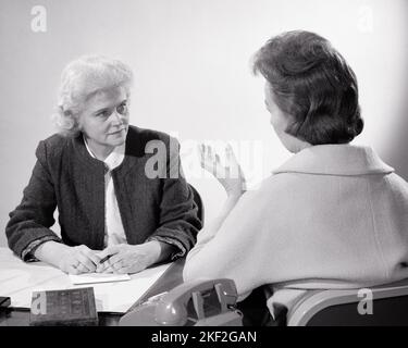 1960s TWO WOMEN COLLEAGUES HAVING A MEETING TALKING ACROSS A DESK BACK VIEW OF ONE WOMAN  - s15055 HAR001 HARS MIDDLE-AGED B&W DESKS MENTOR OCCUPATION MIDDLE-AGED WOMAN ADVISOR CUSTOMER SERVICE NETWORKING CAREERS REAR VIEW OPPORTUNITY OCCUPATIONS MENTAL HEALTH CONNECTION CONCEPTUAL FROM BEHIND ACROSS CONSULTANT SUPPORT COUNSEL COUNSELING ATTORNEY BACK VIEW COOPERATION MID-ADULT MID-ADULT WOMAN SOLUTIONS TOGETHERNESS BLACK AND WHITE CAUCASIAN ETHNICITY COLLEAGUES HAR001 OLD FASHIONED PASTOR Stock Photo