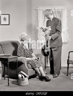 1940s 1950s SENIOR RETIRED COUPLE MAN AND WOMAN AT HOME STANDING HUSBAND PRESENTING BOUQUET OF FLOWERS TO WIFE SITTING IN CHAIR - s3285 HAR001 HARS OLD FASHION RETIRED 1 BOUQUET GIVING STRONG JOY LIFESTYLE CELEBRATION ELDER FEMALES MARRIED RURAL SPOUSE HUSBANDS HOME LIFE COPY SPACE FRIENDSHIP FULL-LENGTH LADIES PERSONS INSPIRATION CARING MALES RETIREMENT SERENITY CONFIDENCE SENIOR MAN SENIOR ADULT B&W PARTNER SENIOR WOMAN RETIREE HAPPINESS OLD AGE OLDSTERS OLDSTER PRESENTING AND EXCITEMENT AT IN OF TO ELDERS CONNECTION CONCEPTUAL STYLISH ELDERLY MAN PERSONAL ATTACHMENT AFFECTION ELDERLY WOMAN Stock Photo