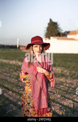 caucasian mature woman with flowered dress pink shawl and big red hat looking at camera in a sown field Stock Photo