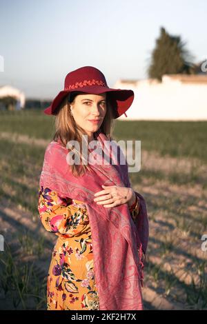 Caucasian mature woman standing in floral dress and pink shawl looking at camera smiling from the orchard on a sunny day Stock Photo
