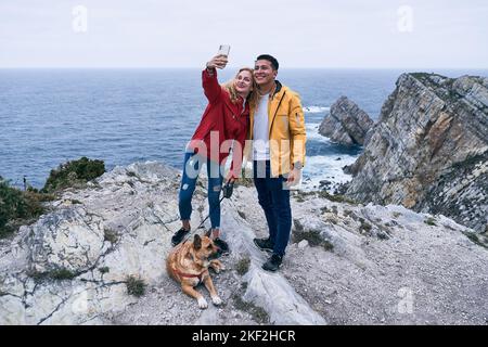 latin boy blond caucasian girl and a big dog lying down smiling while the girl takes a selfie with her smrtphone with her back to the sea, cabo de Stock Photo