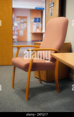 Hospital chair in clinical setting with waiting and reception areas through door in background. Old fashioned hospital chair. Peach and wood. Stock Photo