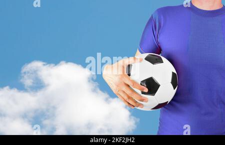 Cropped image of a young man holding a soccer ball with his hand wearing blue tshirt over isolated cloud background with copy space.Online cloud commu Stock Photo