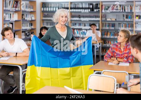 Interesting story about Ukraine while teaching high school students in school library. Teacher holds flag of Ukraine in her hands Stock Photo