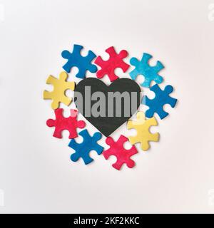 Autism campaign symbols. Colorful puzzle pieces around blackboard stone heart with copy-space. Flat lay, top view, square composition on off white Stock Photo
