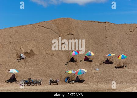 Thai people working together on project in massive sand pile with coloured umbrellas and blue sky background. Stock Photo