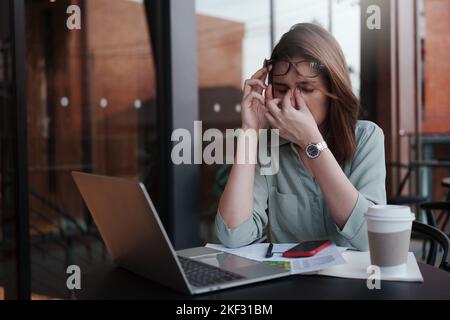 A tired and stressed young woman holds glasses and massages his nose while working hard in a cafe. Stock Photo