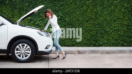 Young woman in a car broke down on the road trying to fix the problem and called a car repair shop or someone close to help. Stock Photo