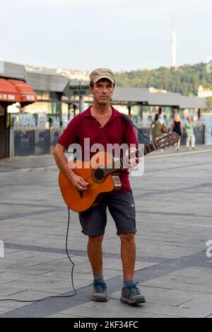 Istanbul, Turkey - August 31, 2022: Male musician Busking playing guitar in Ortakoy district, in front of Bosphorus with Beltas Cafe in the background, and Kucuk Camlica Tower in the far end Stock Photo
