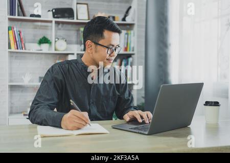 Asian freelance man wearing glasses and smile working typing on laptop and write notes in notebook at home. Entrepreneur man working for business at l Stock Photo