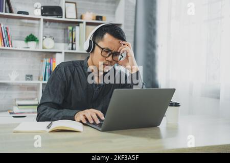 Asian freelance man wear glasses and headphone having stressful depression sad time while working video conferrence meeting on laptop. Depression man Stock Photo