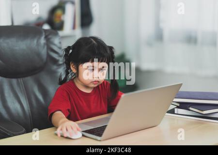 Asian baby girl wearing a red t-shirt use laptop and study online on wood table desk in livingroom at home. Education learning online from home concep Stock Photo