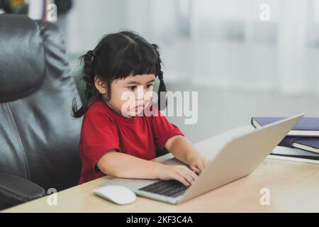 Asian baby girl wearing a red t-shirt use laptop and study online on wood table desk in livingroom at home. Education learning online from home concep Stock Photo