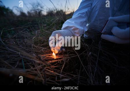 Man burning old dried grass in field. Close up of hand holding burning match and setting fire to dry grass at night. Concept of ecology and human factor in fires. Stock Photo