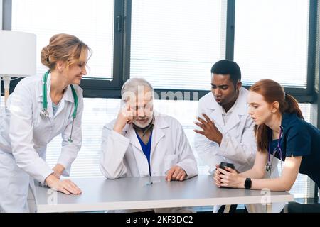 Diverse group of thoughtful multiethnic professional scientist looking at syringe with new vaccine lying on desk and discussing, standing by window. Stock Photo