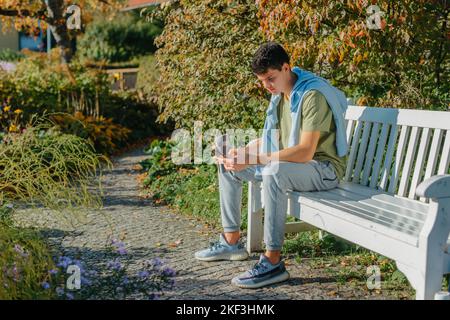 A Teenager Sits On A Bench In The Autumn Park Drinks Coffee From A Thermo Mug And Looks Into A Phone. Portrait Of Handsome Cheerful Guy Sitting On Stock Photo