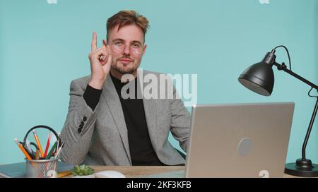 Eureka. Inspired business man sitting at office desk pointing finger up with open mouth, having good idea, plan, startup, showing inspiration motivation gesture, problem solution on blue background Stock Photo