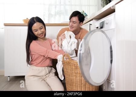 Young Asian married happy couple smiling and doing laundry at home. Boyfriend and girlfriend putting clothes in front loading washing machine together Stock Photo