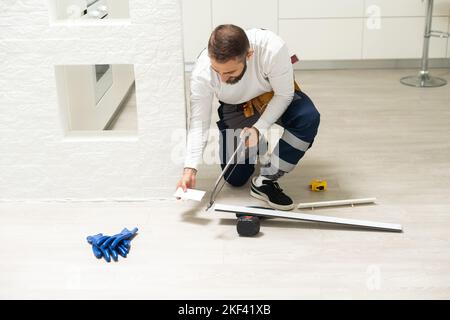 A man installs a floor skirting board. Fixing the plastic skirting board with screws to the wall. Home renovation. Stock Photo